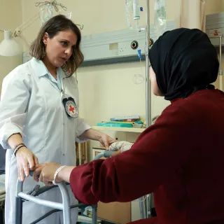 A woman sitting in a hospital bed holds a walker while talking to a female ICRC doctor 
